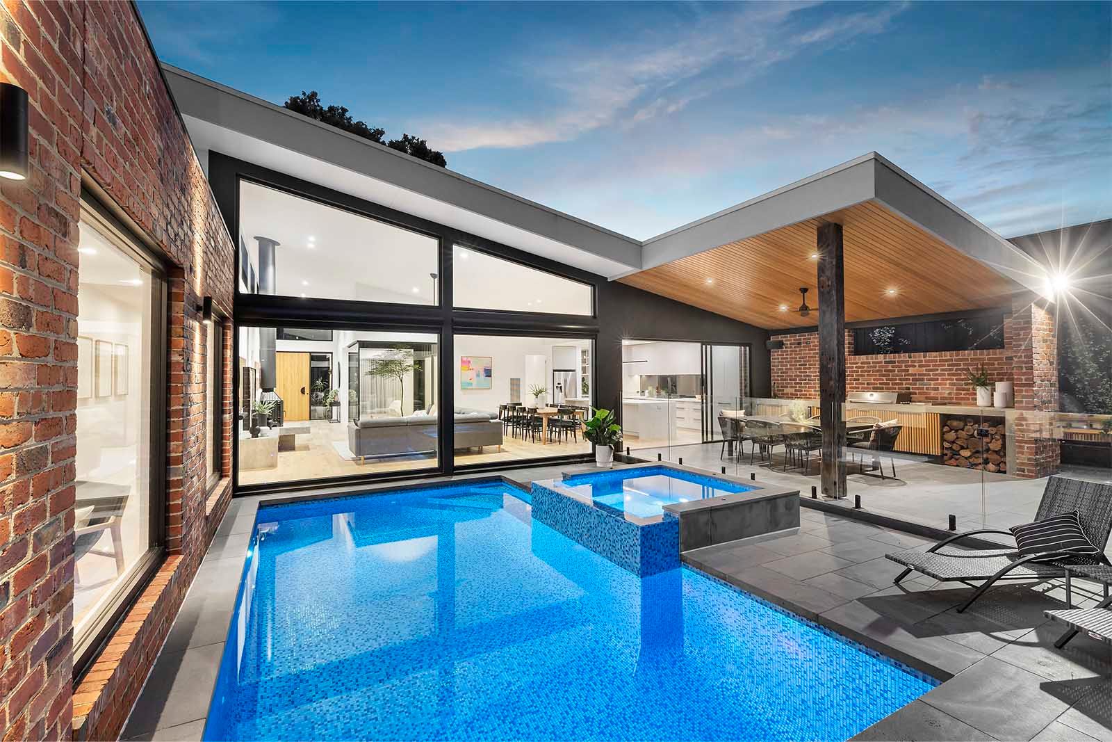 Warranwood home at dusk: pool and spa in the centre, framed by brick, glazing and a timber-lined alfresco